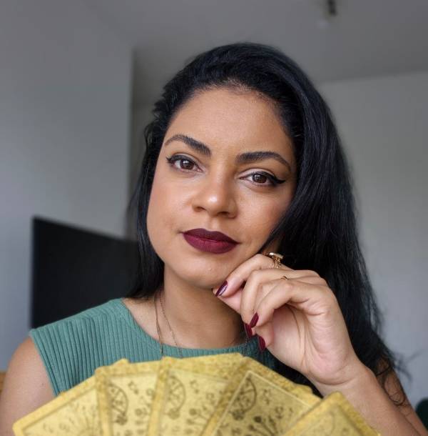 Lane veste verde e segura cartas de tarot douradas, olhando para frente.